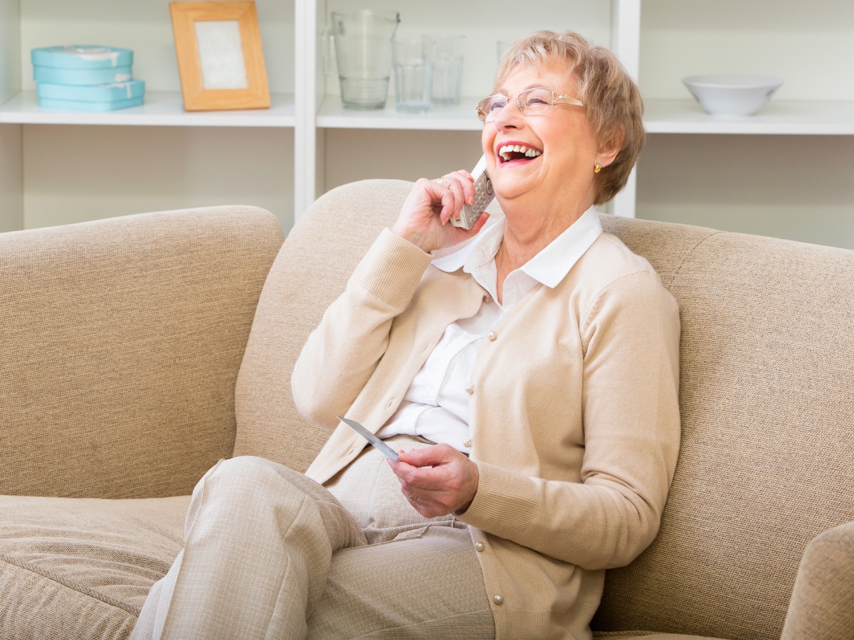 Older woman on a beige sofa, laughing while talking on a cordless phone and holding a card.