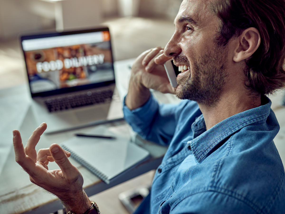 Smiling man on a call at a desk with a laptop displaying a food delivery website in the background, gesturing with his hand.